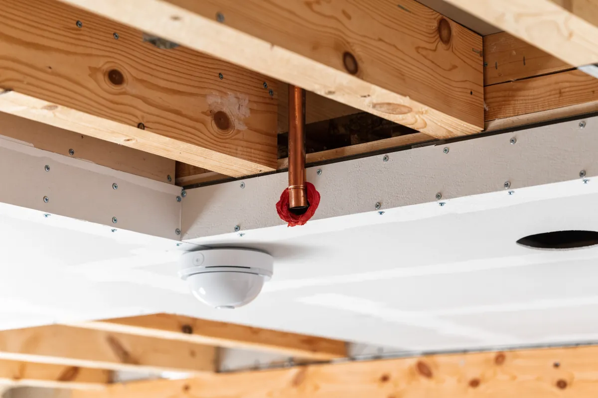 Illustration of fire-rated drywall on a basement ceiling with a pipe penetration sealed with red fire-stop and a smoke alarm, GTA legal suite